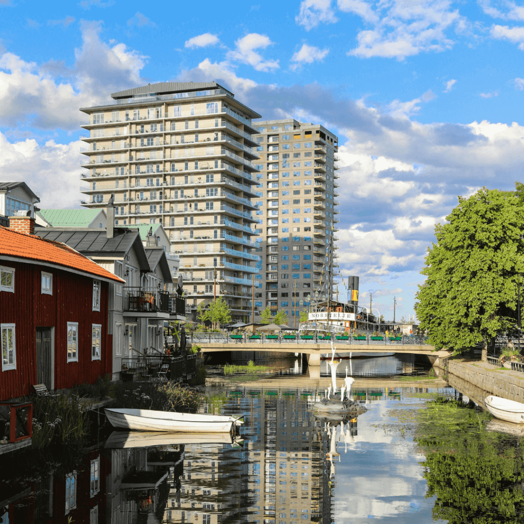 En pittoresk kanalscene i Norrtälje har höga moderna flerbostadshus under en delvis molnig himmel. I förgrunden glider en liten båt förbi en staty av tre svanar. Kanalen, där innovativa webbyråer frodas, kantas av byggnader och grönskande träd.